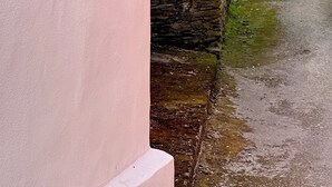 A narrow alleyway with a pink building on one side and a stone wall on the other. A window is visible on the pink building, and there is a metal railing on the stone wall. The alley leads to a street with houses and a view of the sea in the background. - The Cliffs - La Maison du Puits (Porto do Barqueiro)