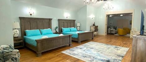 A spacious room with a high ceiling, wooden beams, a large bed with teal bedding, two armchairs, a coffee table, and a chandelier.