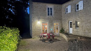 A stone building with a gravel courtyard, a table with chairs, and a well-lit entrance. - Bâtisse du Xixe, Ranville (Ranville)