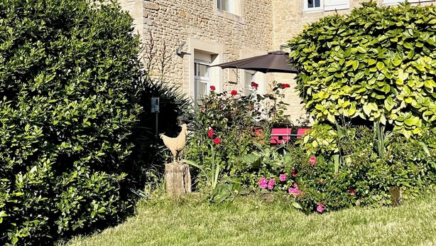 A stone building with a sloped roof, a rooster statue, and a well-maintained garden with various plants and flowers. - Bâtisse du Xixe, Ranville (Ranville)