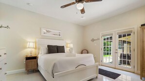 Une chambre avec un lit blanc, des lampes de chevet, un ventilateur de plafond et une vue à travers une porte vitrée.
