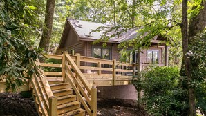 A wooden cabin with a staircase leading to a deck, surrounded by dense forest. - Altitudes Attitude (Blowing Rock)