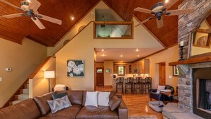 A spacious living room with a brown leather couch, a stone fireplace, and a kitchen in the background. - Crystal Mountain Lodge (Boone)