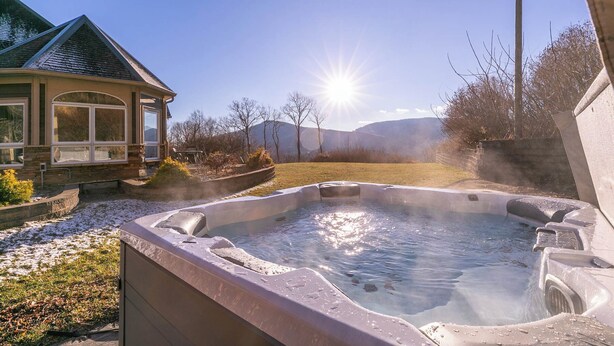 A hot tub with a stone exterior, surrounded by a landscaped garden, a wooden deck, and a house with a green roof. - Mountain Gem (Boone)