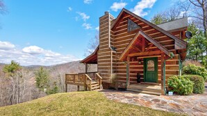 Une cabane en rondins avec une véranda dallée de pierres, une porte verte et un escalier en bois menant à l’entrée.