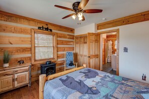 A log cabin bedroom with a bed, a dresser, a window with blinds, a ceiling fan, and a fireplace. - Cozy Bear Cabin (Boone)