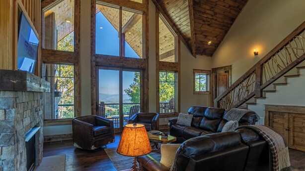 A spacious living room with a stone fireplace, leather furniture, large windows, and a wooden staircase. - Evergreen at Eagles Nest (Elk Park)