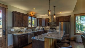 A modern kitchen with dark wood cabinets, a large island, stainless steel appliances, and a view through the windows. - Evergreen at Eagles Nest (Elk Park)