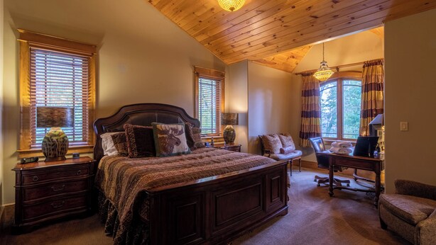 A spacious bedroom with a large bed, wooden headboard, and a chandelier. There are two windows with blinds, a desk with a chair, and a small table with a lamp. - Homestead Lodge at Eagles Nest (Banner Elk)