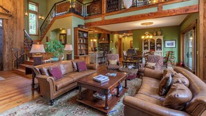 A spacious living area with a high ceiling, wooden beams, a staircase, and a large window. - Homestead Lodge at Eagles Nest (Banner Elk)