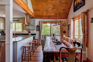 Una cocina rústica con vigas de madera, una larga mesa de madera con taburetes y vistas al verdor exterior.