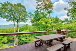 Una terraza de madera con mesas y sillas de picnic, con vista a un bosque frondoso y montañas distantes.