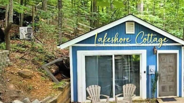 Een klein blauw huisje met een bord waarop staat 'Lakeshore Cottage', twee Adirondack-stoelen, een vuurkorf en een houten terras.