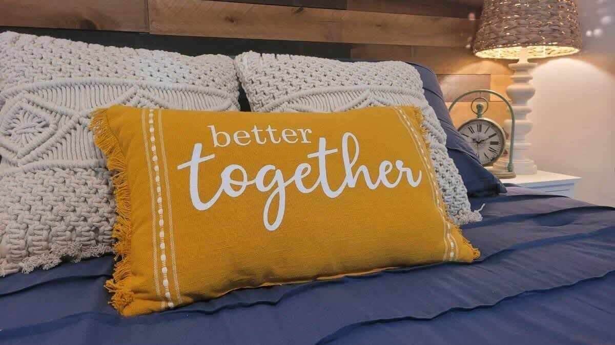 Una cama con colcha azul, una manta tejida blanca y una almohada amarillo mostaza con el texto 'better together'.