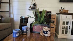 Een gezellige woonkamer met een koffiezetapparaat, twee mokken, een teddybeer, een varen en een vintage koffer.