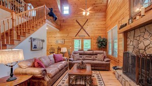 A cozy cabin interior with a wooden staircase, leather sofas, a fireplace, and a large window. - Hebron Falls (Boone)