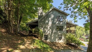 En toetages træhytte med veranda, omgivet af træer og en grusvej.
