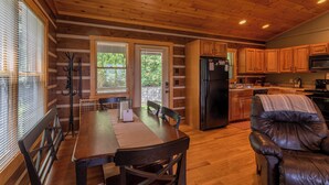 L’intérieur d’une cabane comprend une table à manger en bois, des chaises, un fauteuil inclinable en cuir, un coin cuisine avec des placards en bois et un réfrigérateur.