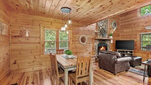 A wooden cabin interior with a dining area, fireplace, leather sofa, and stone fireplace surround. - Among The Trees (Vilas)
