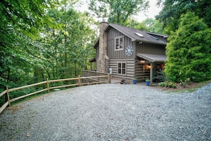 Une cabane rustique avec une cheminée en pierre, une terrasse en bois et une allée gravillonnée.