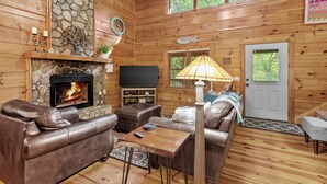 A cozy cabin interior with a stone fireplace, leather sofas, a television, and a lamp. - Among The Trees (Vilas)