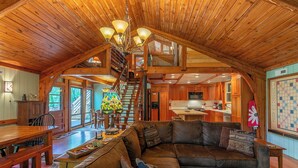 A spacious loft-style room with a wooden ceiling, a dining area with a wooden table and benches, a cozy seating area with a sofa and cushions, and a kitchen with wooden cabinets and a staircase leading to an upper level. - A River Mist (Sugar Grove)