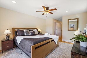 A bedroom with a wooden bed, bedside tables with lamps, a ceiling fan, a mirror, and a potted plant. - House Galini (Morganton)