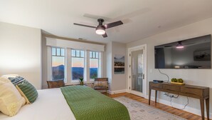 Un dormitorio con una cama, un edredón verde, un escritorio con televisor y vistas a la montaña desde la ventana.