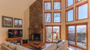 A spacious living room with a stone fireplace, large windows, and a sliding glass door leading to an outdoor deck. - Great Escape at Yonahlossee (Boone)