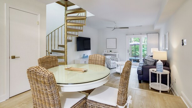 A dining area with a round glass table, wicker chairs, a spiral staircase, and a living room with a sofa and a lamp. - Walk to Shore & Boardwalk: Bethany Beach Retreat (Bethany Beach)