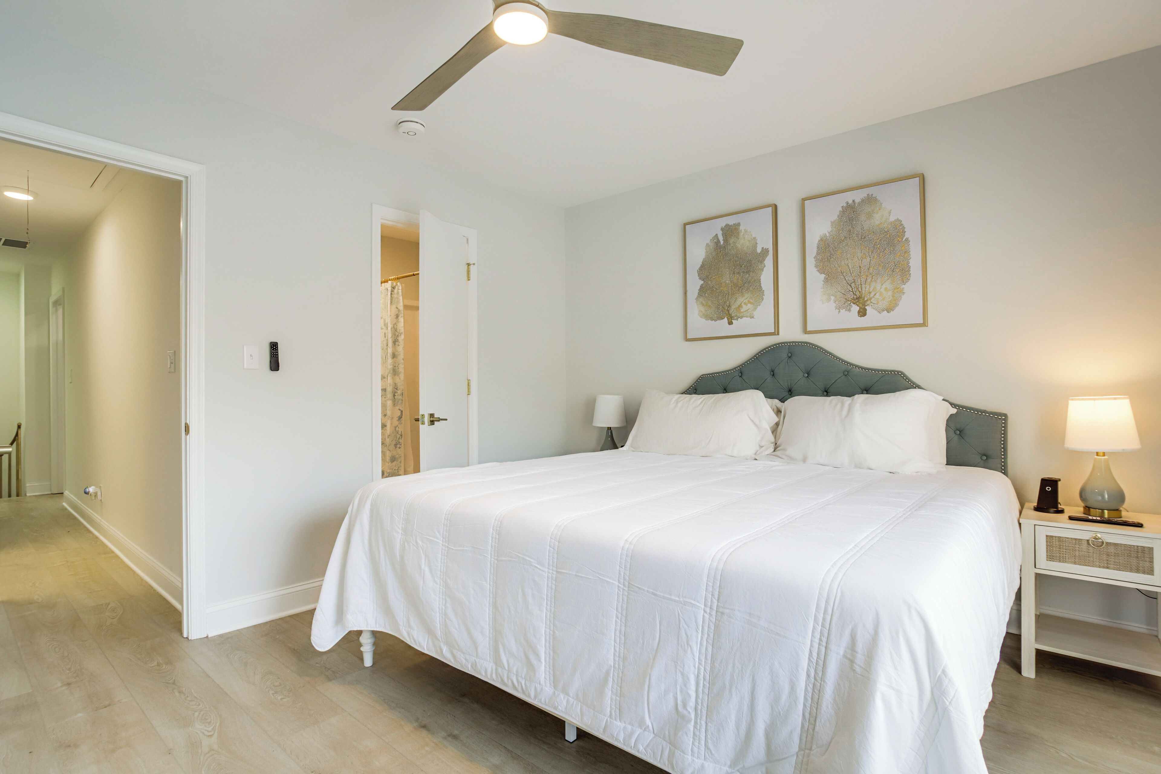 A bedroom with a bed, a ceiling fan, a nightstand with a lamp, and two framed pictures on the wall.