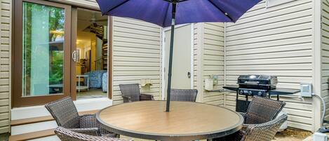 Une terrasse avec une table ronde et des chaises en osier, un barbecue et une porte-fenêtre coulissante donnant sur un espace intérieur.