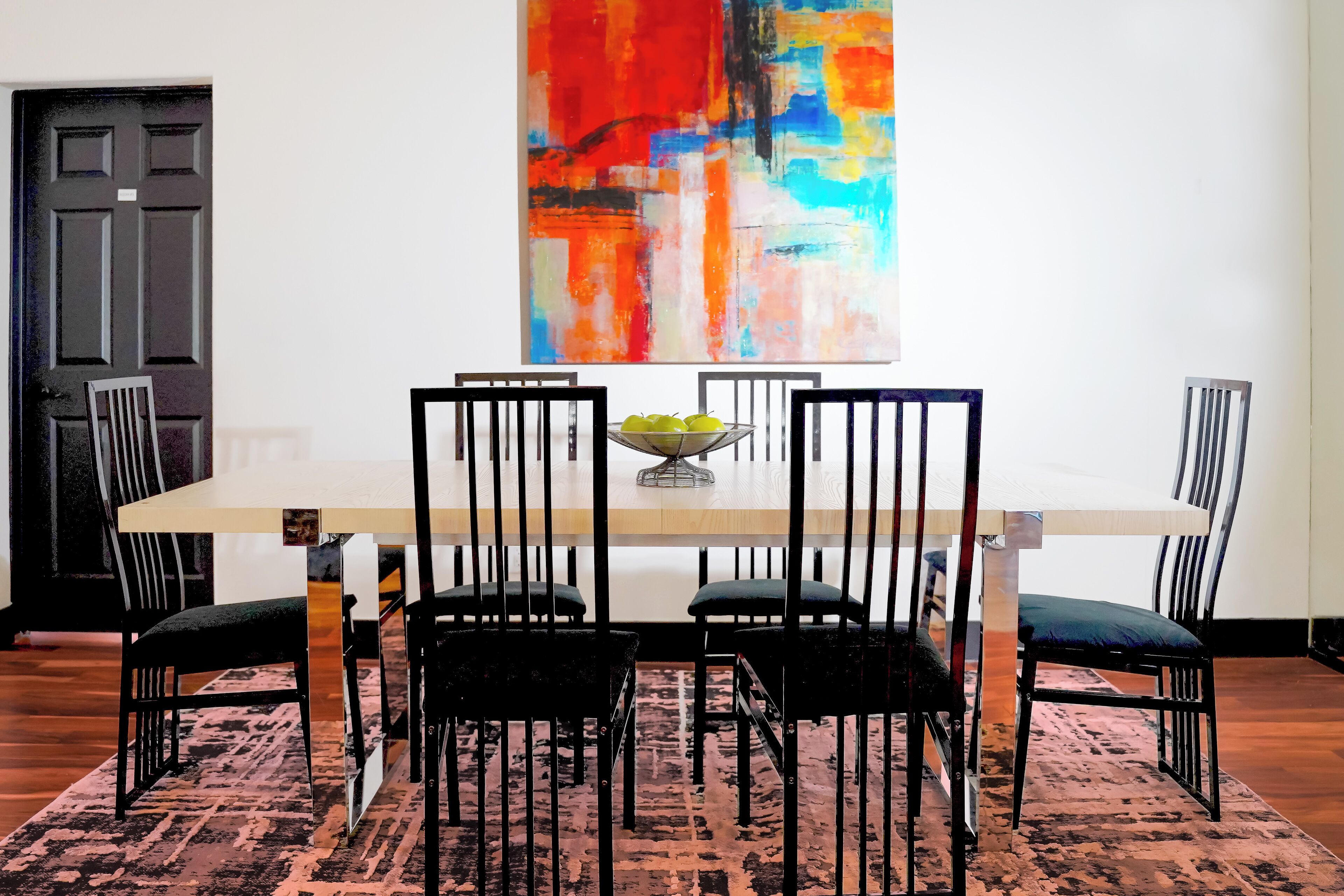 A dining area with a rectangular table, black chairs, and a colorful abstract painting on the wall.