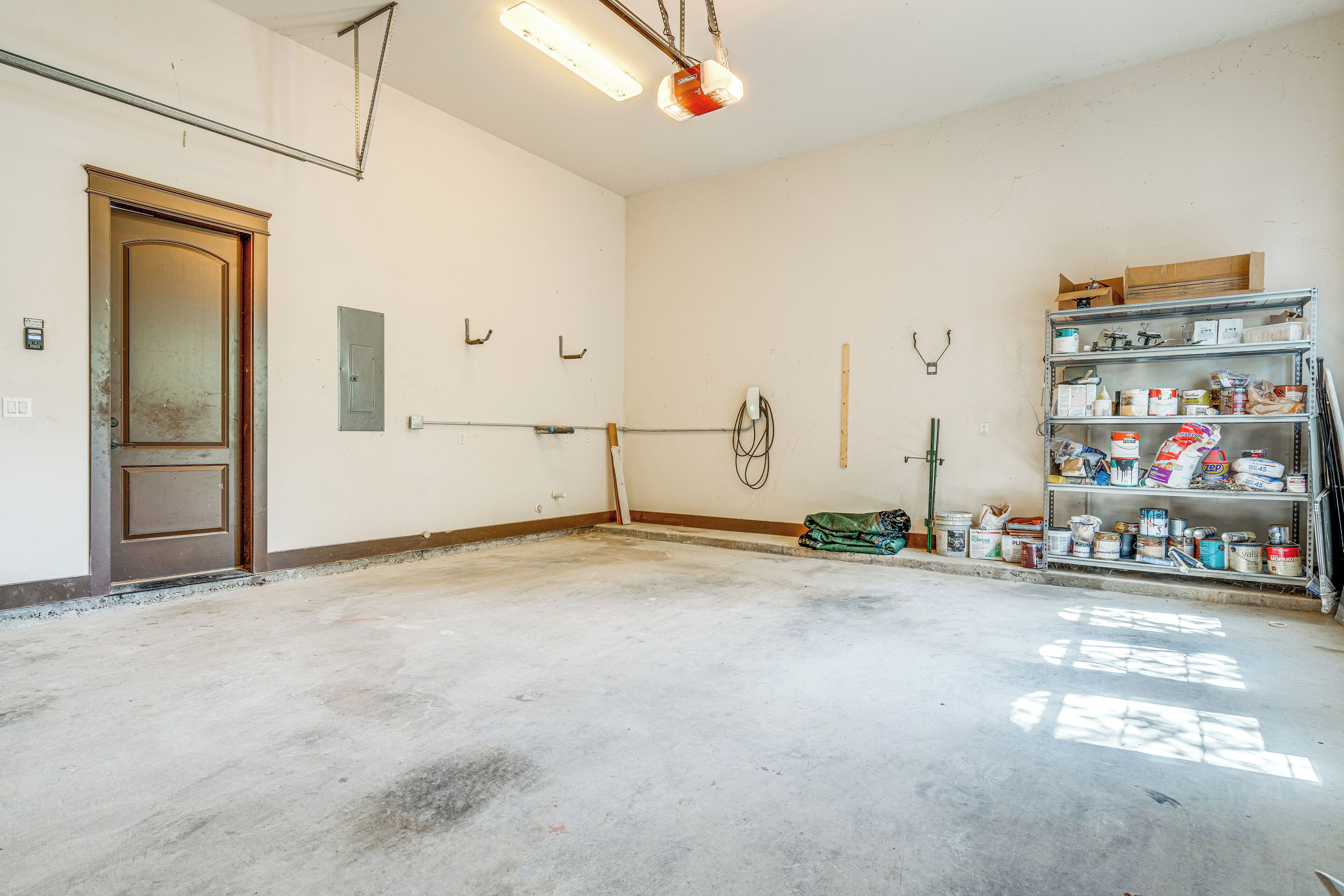 Una habitación amplia con suelo de concreto, una puerta de madera, un estante metálico con varios objetos y una lámpara en el techo.