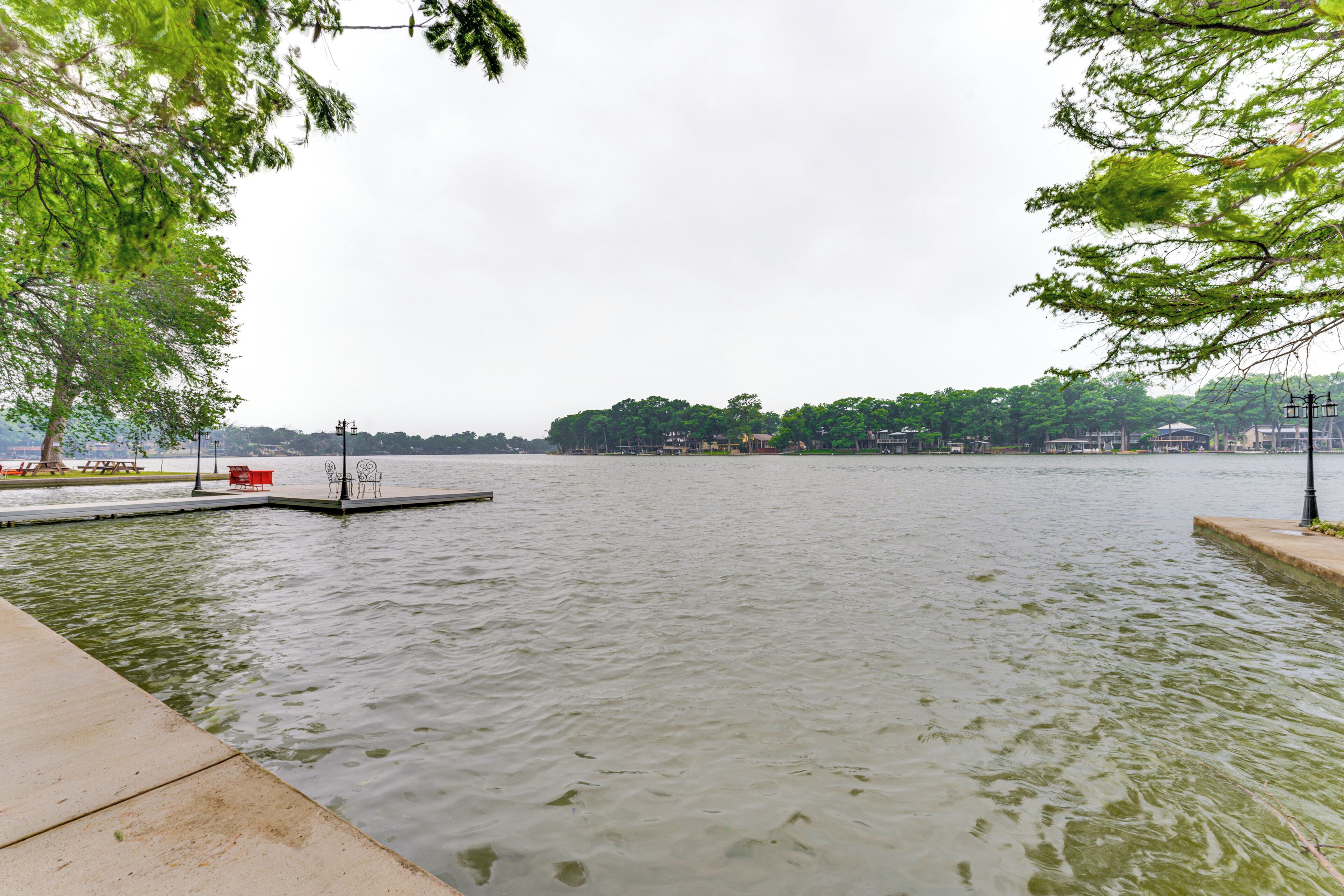 A calm lake with a dock and picnic tables, surrounded by trees and buildings in the distance.