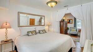 Quarto com cama grande, duas mesas de cabeceira com luminárias, espelho, cadeira e janela com cortinas.
