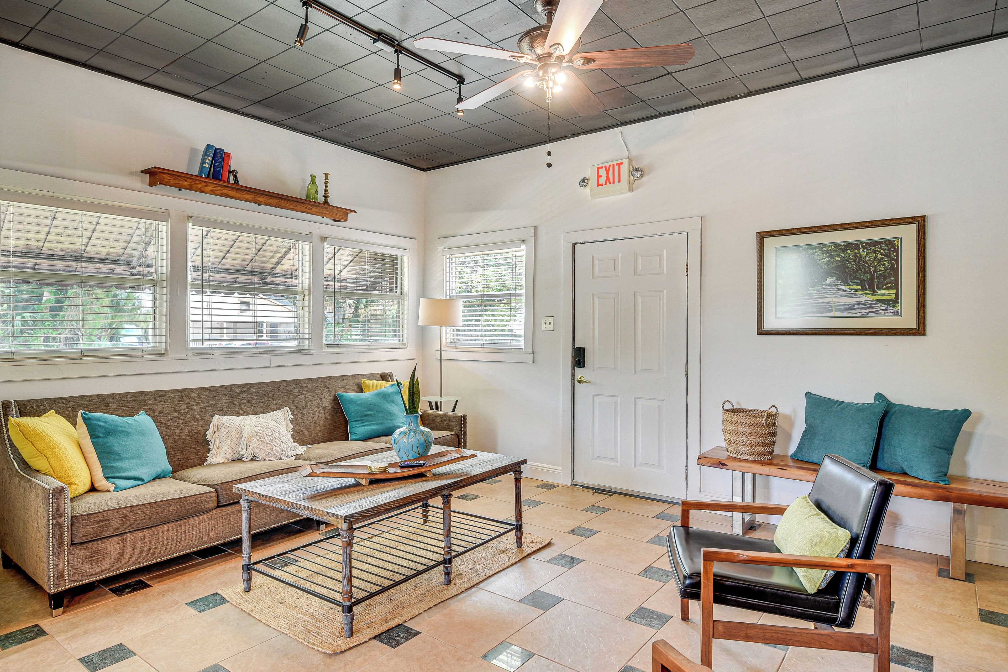 A spacious living area with a sofa, coffee table, and chairs, large windows with blinds, a ceiling fan, and an exit sign.
