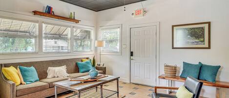 A spacious living area with a sofa, coffee table, and chairs, large windows with blinds, a ceiling fan, and an exit sign.