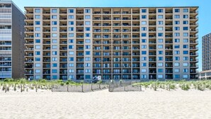 Un hotel de varios pisos con fachada uniforme, balcones y una playa arenosa al frente.