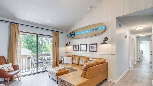 A spacious living room with a large tan sectional sofa, a coffee table, and a dining area with chairs. A surfboard decoration hangs on the wall, and there are framed pictures above the sofa. - Amelia Island Condo w/ Pool Access: Walk to Beach! (Fernandina Beach)