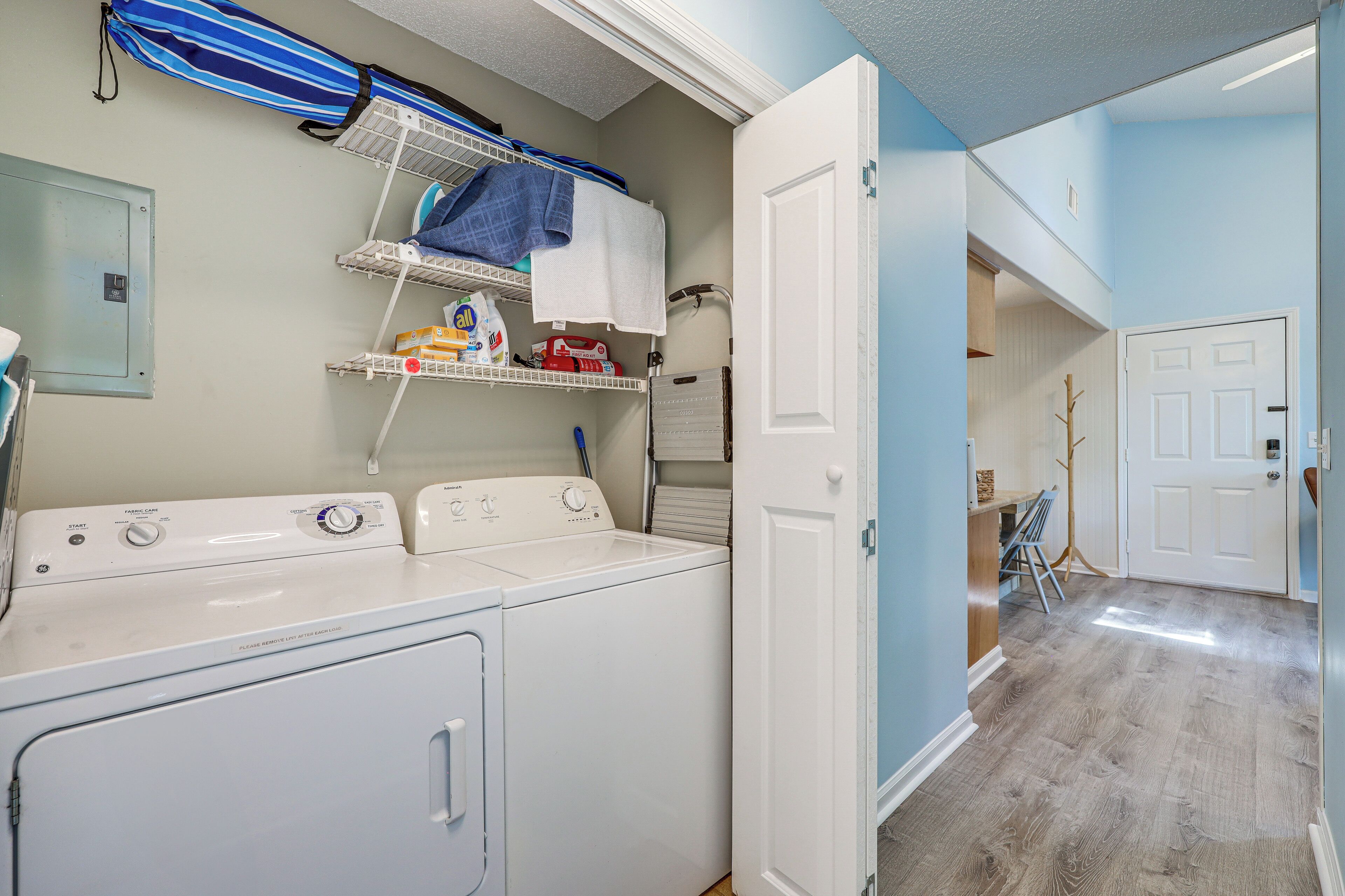 A laundry area with a front-loading washer and dryer, shelves with laundry supplies, and a door leading to another room.