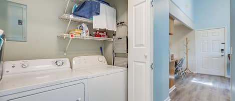 A laundry area with a front-loading washer and dryer, shelves with laundry supplies, and a door leading to another room.