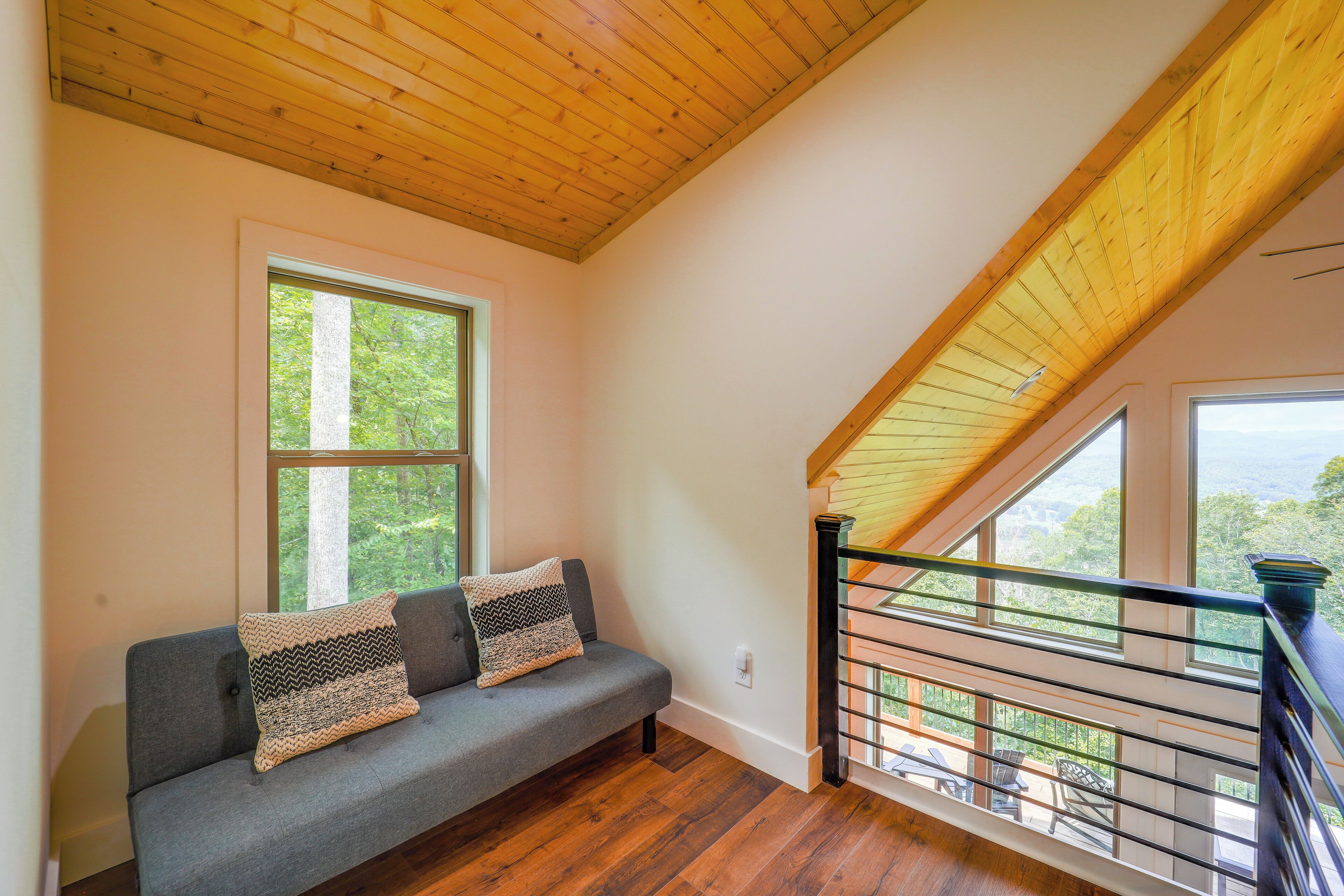 A room with a wooden ceiling, a grey sofa with patterned cushions, and a view of trees through large windows.