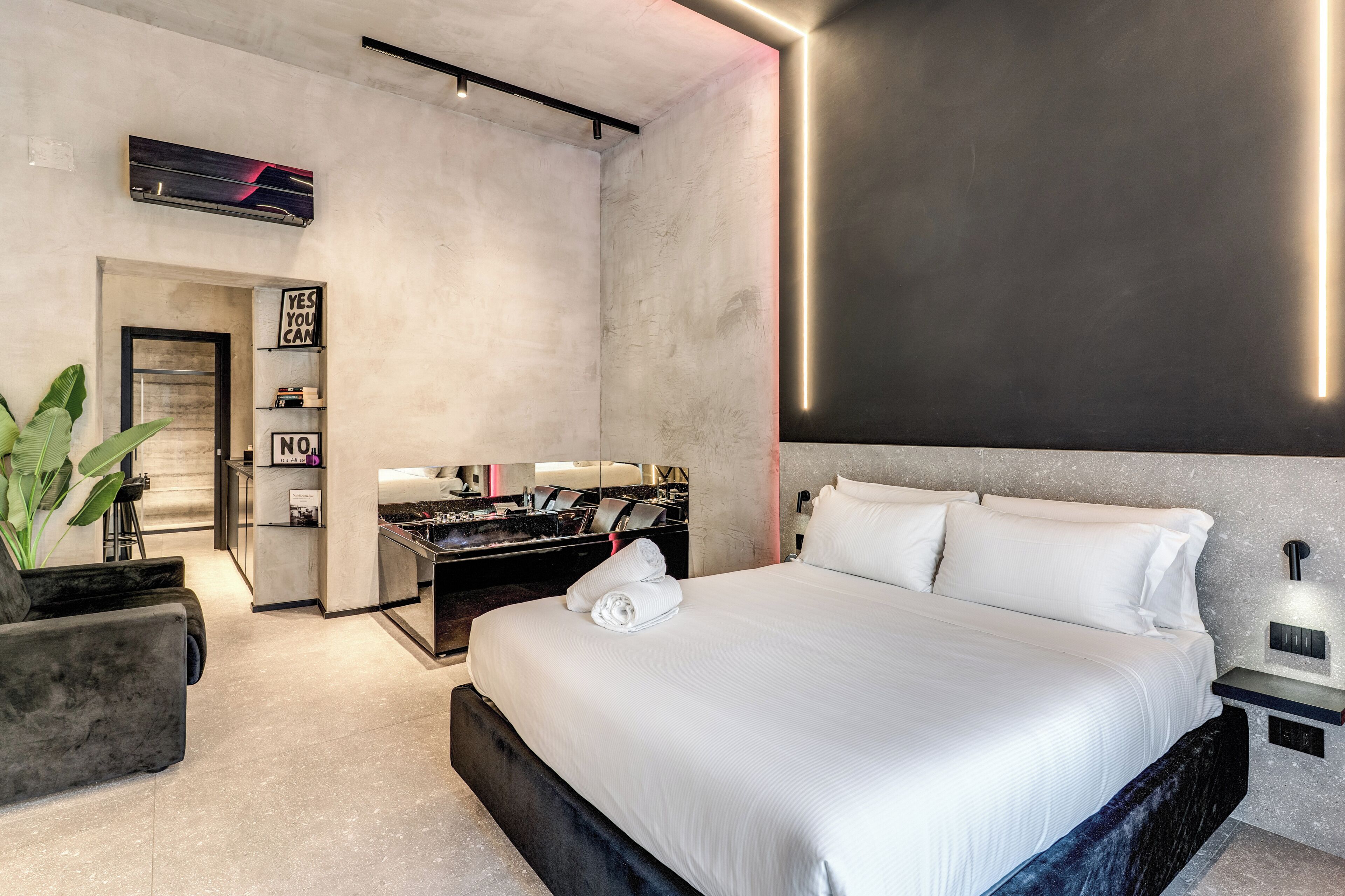 A modern hotel room with a large bed, a desk with a chair, a potted plant, and a mirror.