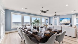 A spacious dining area with a long wooden table set for six, surrounded by white chairs. There are two ceiling fans, a large window with a view of the ocean, and a television mounted on the wall. - Bluebell: Beachfront, Private Pool, Arcade Games, Plantation (St. George Island)
