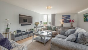 A modern living room with a flat-screen TV, a glass coffee table, grey sofas, a grey entertainment unit, and a large window with curtains. - Westburn Lane Apartments (No 2) (St Andrews)