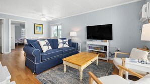 A living room with a blue sofa, wooden coffee table, and white armchairs. There is a flat-screen TV mounted on the wall and a white cabinet with baskets. - Ocean Block 2 BR Unit Ready for Summer Fun! (Ocean City)