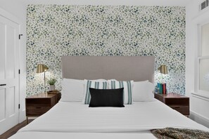 A bedroom with a bed, a headboard, two bedside tables with lamps, and a patterned wallpaper.