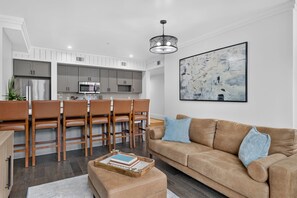 A modern living room with a brown leather sofa, a coffee table with books, and a dining area with wooden chairs and a kitchen in the background.