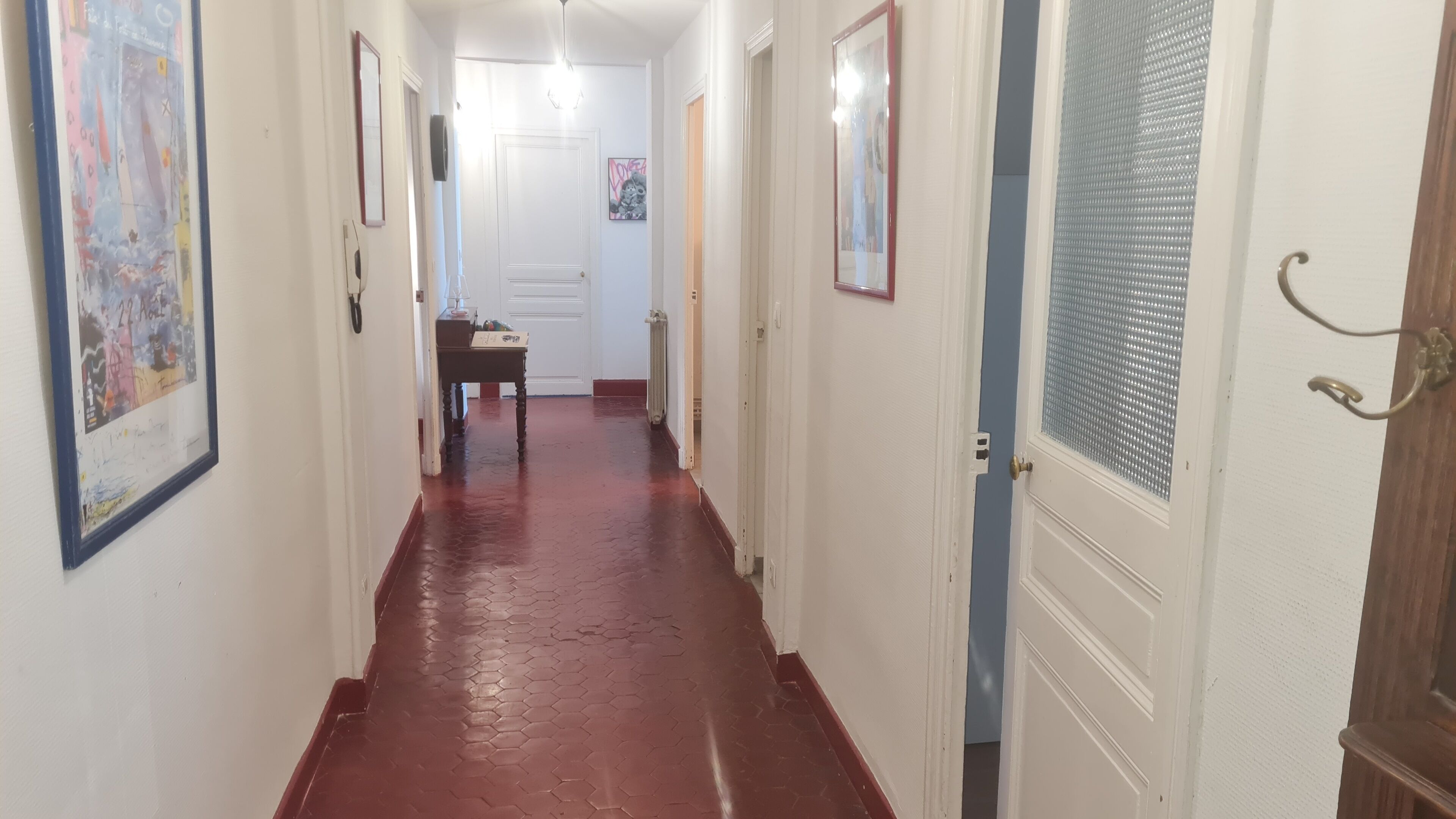 A hallway with a red tiled floor, white walls, and doors leading to other rooms. There are framed pictures on the walls and a table with a lamp in the distance.